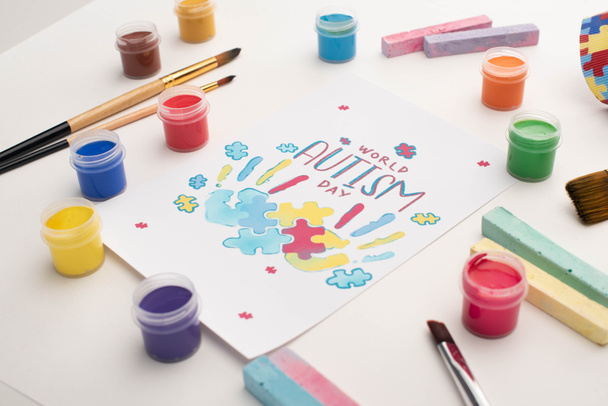 kaart met Wereld Autisme Dag belettering en schilderen van puzzel en handafdrukken op wit met verf borstels, krijt en verf - Foto, afbeelding