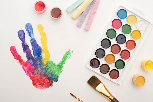 κορυφαία άποψη πολύχρωμο αποτύπωμα χεριού, κιμωλίες, πινέλα και χρώματα σε λευκό χρώμα για την Παγκόσμια Ημέρα Ευαισθητοποίησης του Αυτισμού - Φωτογραφία, εικόνα