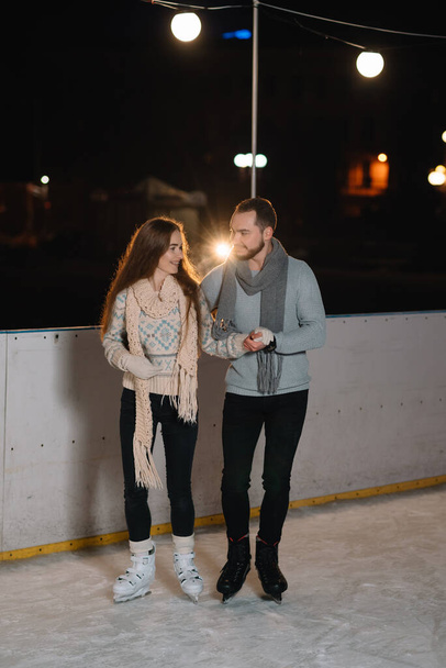 Couple on the city rink in a winter evening. Guy helping nice girl to skate on the ice in the dark night and twinkles lighting above them - Photo, Image