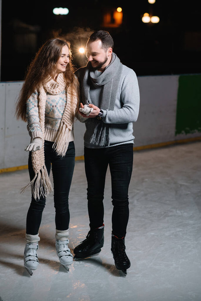 Couple on the city rink in a winter evening. Guy helping nice girl to skate on the ice in the dark night and twinkles lighting above them - Foto, Imagen