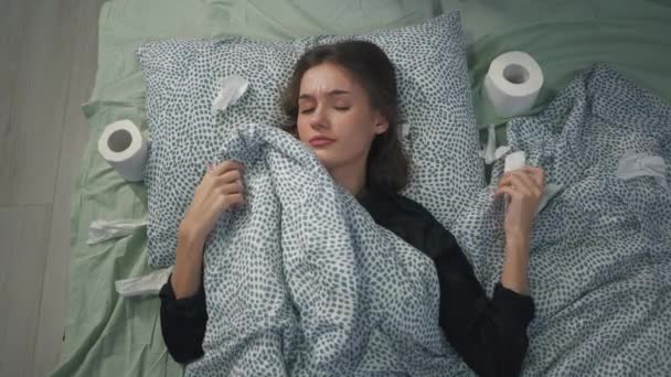 The sick girl lies in bed under the covers and blows her nose. Toilet paper in a coronavirus pandemic like a handkerchief. Coronavirus protection. Home treatment at the time of carntin. - Materiał filmowy, wideo