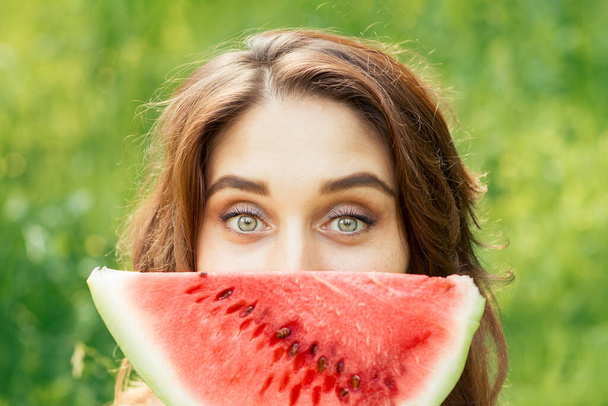 Krásná mladá žena drží plátek melounu před obličejem na zeleném přírodním pozadí. Dívka drží plátek melounu blízko obličeje zblízka. - Fotografie, Obrázek