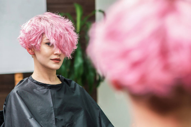 Девушка с розовыми волосами смотрит в зеркало и ждет парикмахера в парикмахерской красоты
 - Фото, изображение