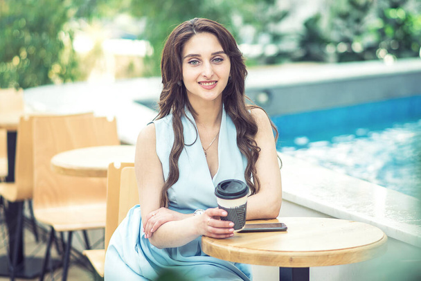 Jovem mulher bonita está segurando caneca de papel em um café enquanto olha para a câmera ao ar livre. xícara de café na mão mulher
. - Foto, Imagem