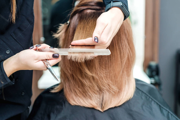 Les mains féminines du coiffeur tiennent les cheveux roux de la femme avec un peigne entre les doigts dans le salon de beauté gros plan. - Photo, image