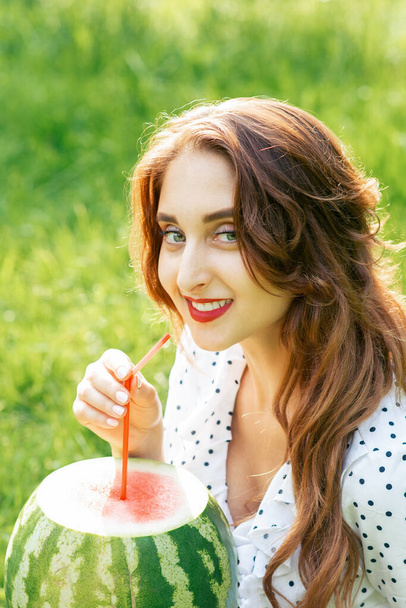 Frau in weißem Kleid hält Wassermelone mit Cocktailstroh auf Grashintergrund. Adlt Mädchen hält eine ganze Wassermelone aus nächster Nähe. - Foto, Bild