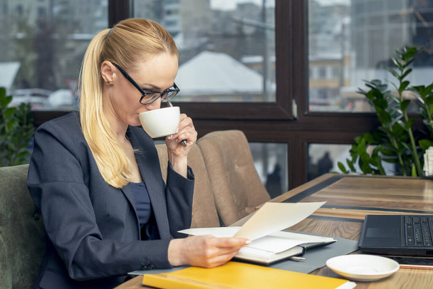 Kahve dükkanında oturan gözlüklü genç bir iş kadını laptopun önünde dokümanları okuyor ve kahve içiyor. İş konsepti. - Fotoğraf, Görsel