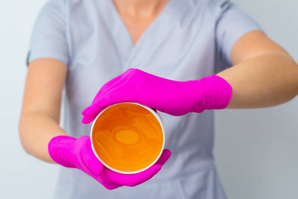 Kosmetikerin in rosa Handschuhen hält Glas mit flüssiger gelber Paste für Epilation, Enthaarung und Schönheitskonzept, Nahaufnahme. - Foto, Bild