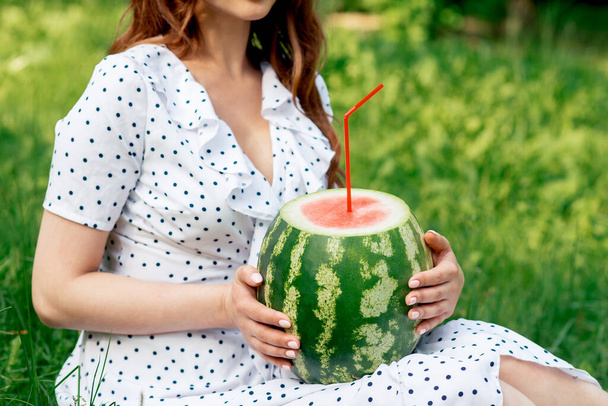Woman is holding a whole watermelon with cocktail straw on grass background. Girl holds a whole watermelon. - Fotografie, Obrázek