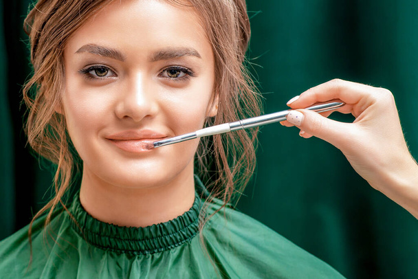 Mano de artista de maquillaje aplica lápiz labial rosa para mujer hermosa joven, de cerca
. - Foto, Imagen