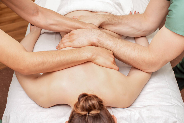 Κοντινό πλάνο γυναίκα λαμβάνει μασάζ στην πλάτη σε τέσσερα χέρια από δύο άνδρες θεραπευτές. - Φωτογραφία, εικόνα