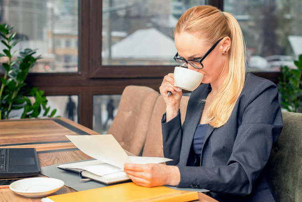 Молодая деловая женщина в очках, сидящая за столом в кафе, читает документы перед ноутбуком и пьет кофе. Концепция бизнеса
. - Фото, изображение