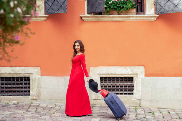 Krásná cestovatelka se prochází po ulici s kufrem. Cestovatel mladá žena v dlouhých červených šatech procházky ve městě ulici s kufrem. - Fotografie, Obrázek