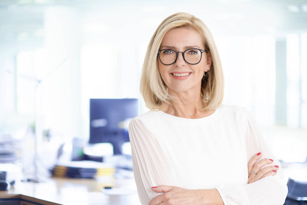Portrait shot of attractive senior businesswoman standing with arms crossed in the office while looking at camera and smiling. - Foto, Imagem