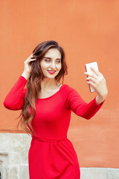 Beautiful adult girl is making selfie by smartphone on orange background. Fashionable nice girl taking selfie on background of orange wall. - Foto, immagini