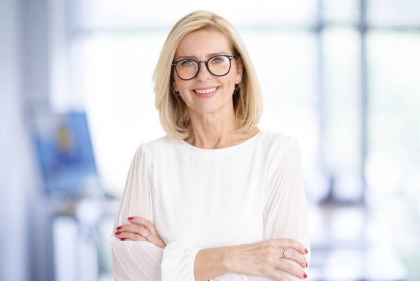 Portrait shot of attractive senior businesswoman standing with arms crossed in the office while looking at camera and smiling. - Fotografie, Obrázek