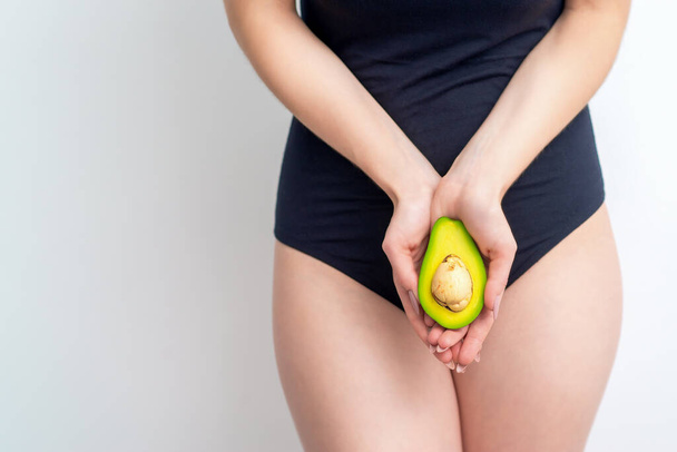 Half an avocado in hands of young female covering bikini area on white background with copy space. Concept of the health of the intimate organs. - Photo, Image