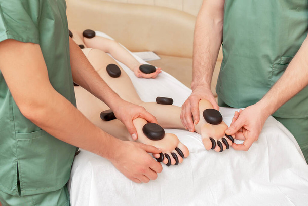 Kobieta otrzymująca masaż gorącymi kamieniami na nogach w czterech rękach przez dwóch masażystów. - Zdjęcie, obraz