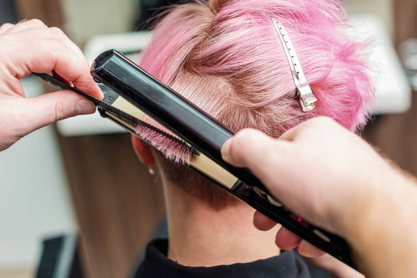 professional hairdresser straightening pink hair with hair straighteners in beauty salon, concept barber salon, female stylist - Photo, Image