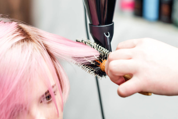 A fodrász rózsaszín hajat szárít hajszárítóval és kerek kefével. Fodrász hajszárító rózsaszín nő haja közelről. - Fotó, kép