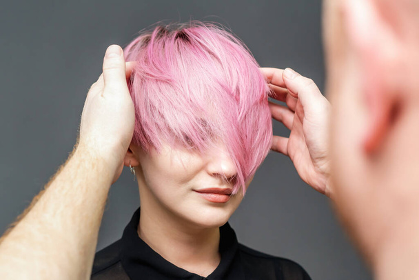 kapper is het controleren van korte roze kapsel van jonge vrouw op grijze achtergrond, close-up. - Foto, afbeelding