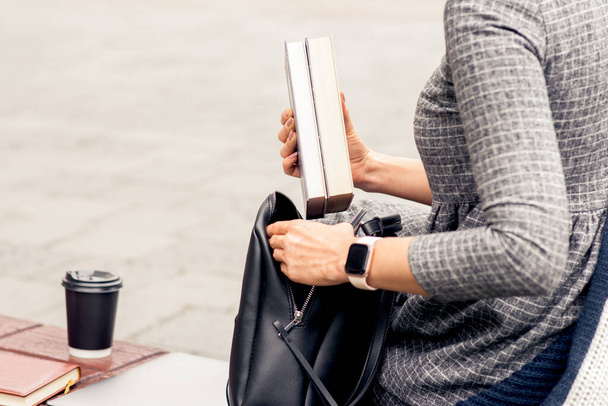 Female student puts books into into black backpack close up. Hands of woman student is putting books into backpack outdoors. Student life concept. - 写真・画像