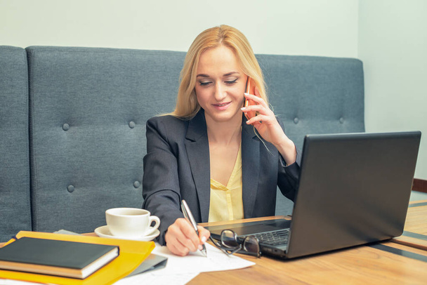 bastante joven mujer de negocios está hablando por teléfono mientras escribe en un papel en la oficina, concepto de negocio
 - Foto, imagen