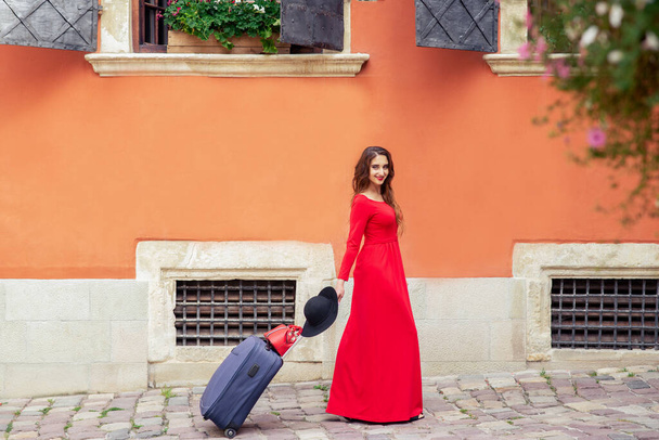 Mulher viajante bonita puxa a mala na rua da cidade. Viajante jovem mulher em vestido vermelho longo caminha na rua da cidade com mala
. - Foto, Imagem