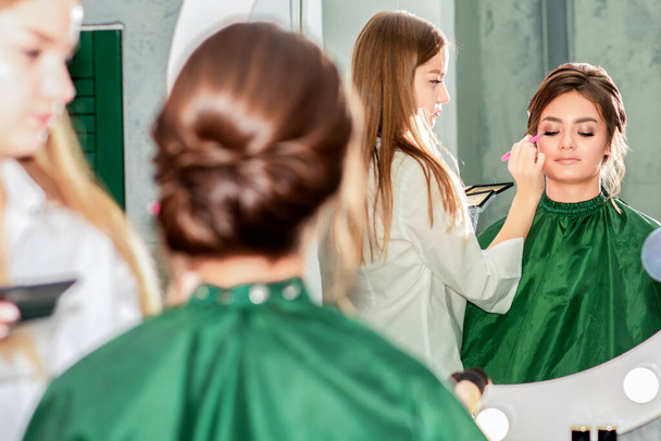 Maquillaje artista haciendo maquillaje profesional para mujer joven en espejo reflexión en salón de belleza
. - Foto, imagen