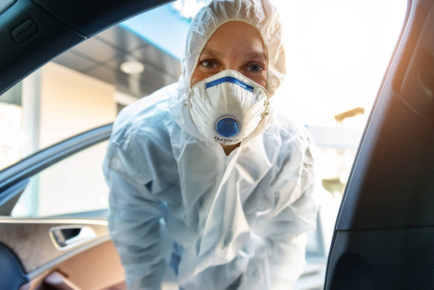 Πορτρέτο της κουρασμένης εξαντλημένης γυναικείας γιατρού, επιστήμονα ή νοσηλευτή που φοράει μάσκα προσώπου και προστατευτική στολή βιολογικού κινδύνου ανοιχτή πόρτα αυτοκινήτου σε υπαίθριο δρόμο. Κίνδυνος επιδημίας του ιού του Coronavirus covid-19. - Φωτογραφία, εικόνα