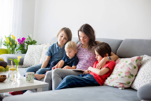 Young woman, mother with three kids, reading a book at home, hugging and laughing, family happiness - 写真・画像