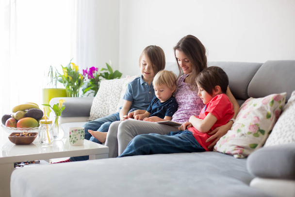 Молода жінка, мати з трьома дітьми, читає книгу вдома, обіймає і сміється, сімейне щастя
 - Фото, зображення