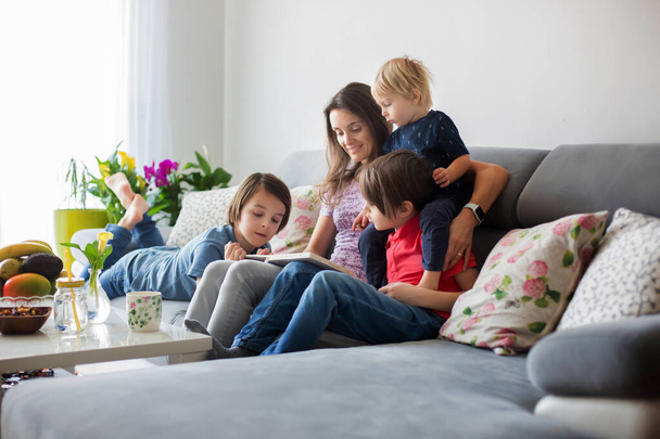 Young woman, mother with three kids, reading a book at home, hugging and laughing, family happiness - Photo, Image