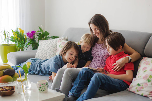 Young woman, mother with three kids, reading a book at home, hugging and laughing, family happiness - Foto, Imagem