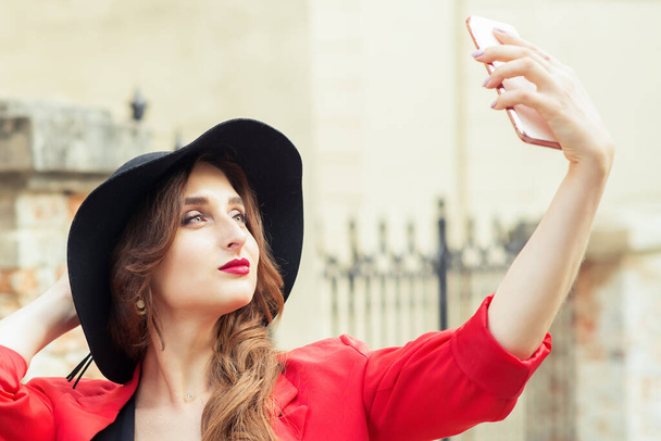 Beautiful girl in black hat makes selfie herself at city street. - Фото, зображення