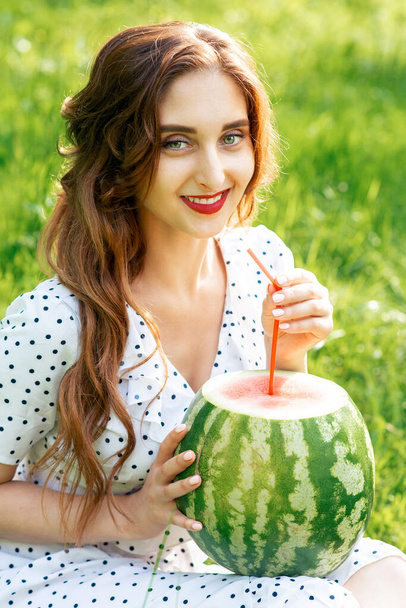Portrait of beautiful young woman holds watermelon siting on grass outdoors. - Фото, изображение