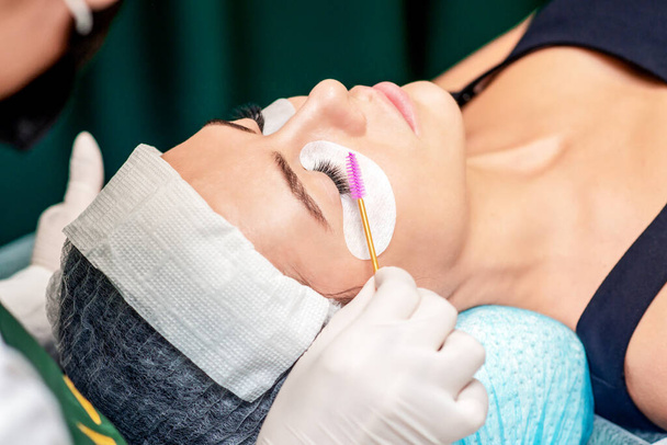 Cosmetologist uses eyelash brush for eyelashes during eyelash extension procedure in a beauty salon. - Photo, Image