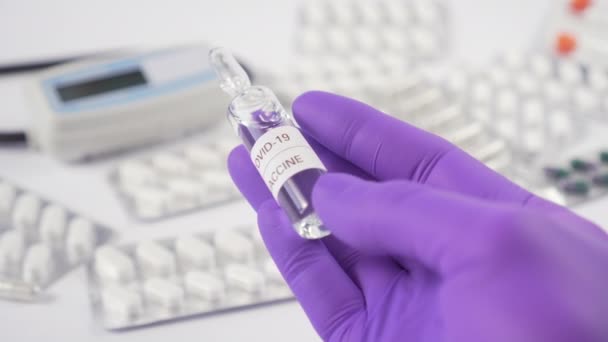 Φύσιγγα εμβολίου του κερατοειδούς στο χέρι με μπλε προστατευτικά γάντια πάνω από ένα λευκό τραπέζι με ιατρικό εξοπλισμό και δισκία. Αναζητήστε ένα φάρμακο για το COVID-19 - Πλάνα, βίντεο