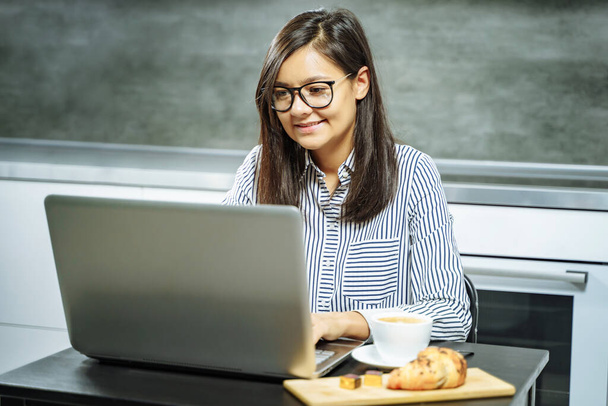 Smiling business woman with glasses working on a laptop in the kitchen at home. - 写真・画像