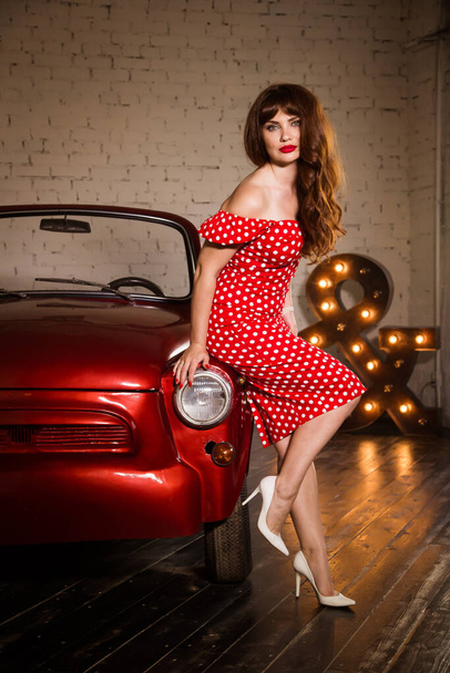 Beautiful woman in a tight dress, dressed in pin-up style. Caucasian brunette model posing in retro fashion and standing at the old car without a roof. Red lips. - Fotografie, Obrázek
