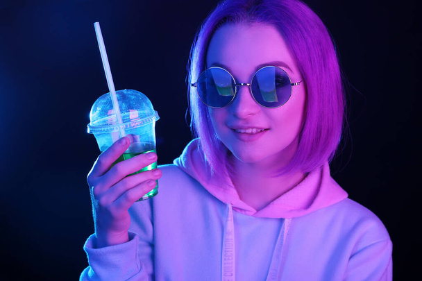 Woman in neon light and sunglasses. In the club and holds in his hand a glass with a drink, a cocktail. Night life. Generation Y, millennials. - Photo, Image