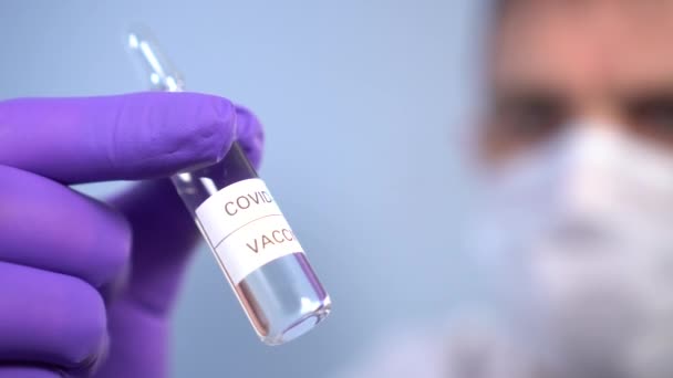 Coronavirus vaccine ampoule in doctor's hands in face mask and protective medical gloves on a blue background. Testing and use of a new drug from COVID-19 - Footage, Video