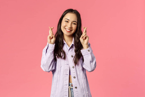 Portrait of hopeful, optimistic happy girl hold fingers crossed for dream come true, making wish, smiling and looking camera, having faith in all be good, pink background - Photo, Image