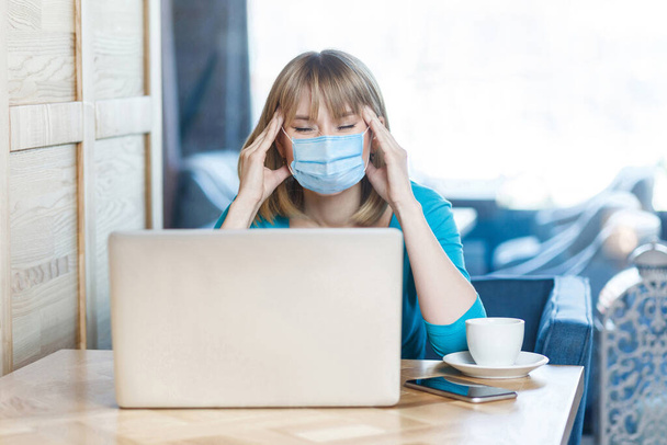 Headache or confusion. Portrait of young woman with surgical medical mask is sitting and working on laptop and holding her painful head or thinking. Indoor working, medicine and health care concept. - Foto, afbeelding