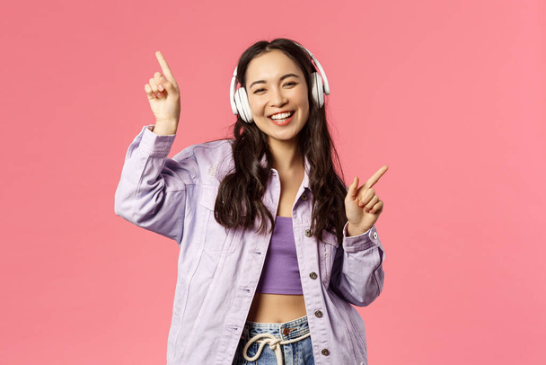 Lifestyle, technology and people concept. Portrait of carefree pretty young asian girl having fun, dancing joyfully, listening music in eadphones, raising hands up, singing along, pink background - Foto, Imagem