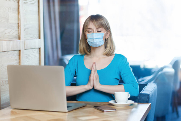 Portrait de jeune femme blonde calme avec masque médical chirurgical en t-shirt bleu est assis et travaille sur ordinateur portable et essayer de se détendre dans la pose de yoga. Concept de travail intérieur, médecine et soins de santé
. - Photo, image