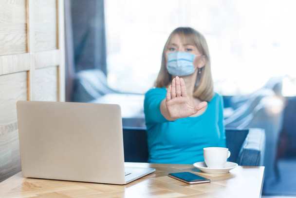 Stop, don't come any closer. Portrait of serious young woman with surgical medical mask sitting and showing stop with hands and warning about distance. Indoor working, medicine and health care concept - Foto, Imagem