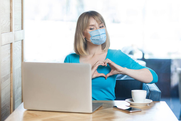 I love you. Portrait of young woman with surgical medical mask sitting and looking at laptop display with heart gesture on video call. Indoor distance working, medicine and health care concept. - Photo, Image