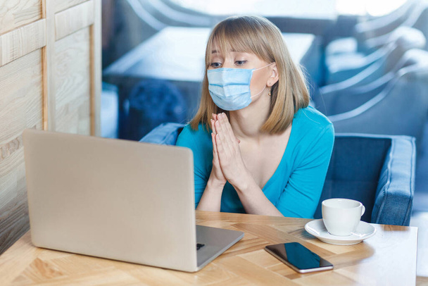 please help. Portrait of worry young woman with surgical medical mask is sitting and working on laptop and looking display and begging on video call. Indoor working, medicine and health care concept. - Foto, Bild
