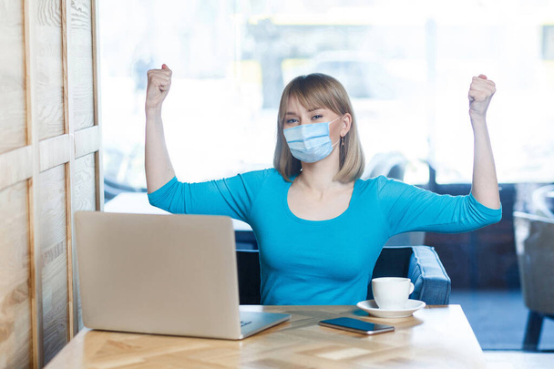 I am strong. Portrait of young woman with surgical medical mask in blue t-shirt is sitting and looking at camera and showing her biceps and satisfied. Indoor working, medicine and health care concept. - Photo, Image